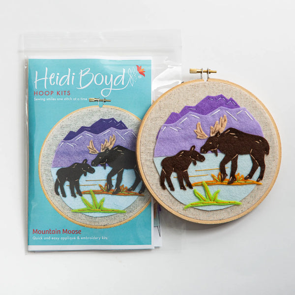 Mountain moose hoop kit in clear plastic package next to completed example with two moose and purple mountains